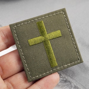 May include: Olive green embroidered patch with a lime green cross design. The square patch has a stitched border with white thread. The patch is made of a textured fabric.