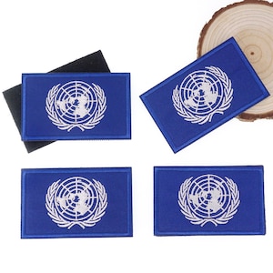 May include: Four blue rectangular patches with the United Nations emblem embroidered in white. The emblem features a world map encircled by olive branches. One patch has a black velcro backing.