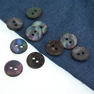 May include: A selection of round buttons, each with two holes. The buttons display a dark, iridescent sheen with green, purple, and brown hues. They are arranged on a blue fabric background.