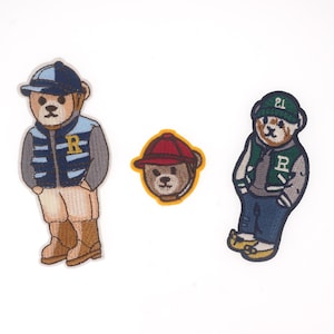 Puede incluir: Tres parches bordados para planchar que representan osos de dibujos animados con diferentes atuendos. El primer oso lleva una chaqueta a rayas azul y blanca, una gorra de béisbol azul y botas marrones. El segundo oso lleva una gorra de béisbol roja. El tercer oso lleva una chaqueta verde y gris, una gorra de béisbol verde y pantalones vaqueros azules.