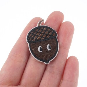 May include: A brown and white embroidered acorn with a smiling face. The acorn is on a white background.