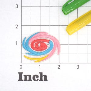 May include: A colorful embroidered patch in the shape of a spiral. The patch is made up of four layers of fabric in pink, red, blue, and yellow.
