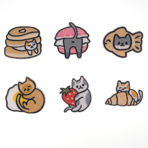 May include: Set of 7 iron-on embroidered patches featuring cute cats in various scenes. The patches include a cat in a pancake stack, a cat in a macaron, a cat in a fish-shaped pastry, a cat in an egg, a cat holding a strawberry, and a cat in a croissant.