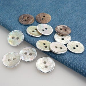 May include: A selection of decorative buttons in different styles. Some buttons are round with a pearlescent finish and snowflake designs. Others are brown and textured. All buttons have two holes for sewing.