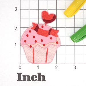 Może przedstawiać: R&oacute;żowy cupcake z czerwono-białym lukrem, czerwonymi posypkami i czerwonym sercem na g&oacute;rze. Cupcake ma około 5 cm szerokości.