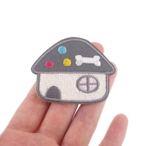 May include: A gray and white embroidered iron-on patch featuring a cartoon house with a bone on the roof and colorful dots. The house has a window and a door.