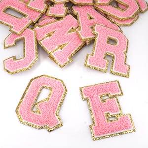 Op de afbeelding: Roze vilt letters met gouden glitterrand. De letters zijn in hoofdletters en omvatten de letters J, A, R, Q en E.