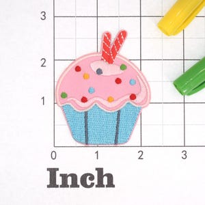 Może przedstawiać: R&oacute;żowy cupcake z niebieskim lukrem i kolorowymi posypkami. Na wierzchu cupcake'a znajduje się czerwony i biały cukierek. Cupcake ma około 5 cm szerokości.