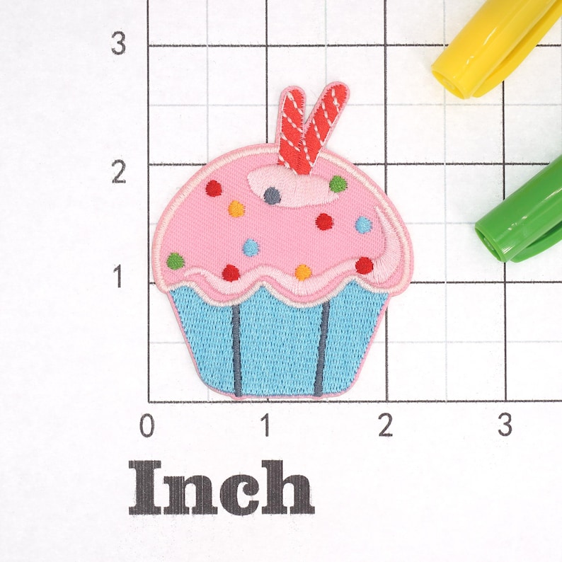 Może przedstawiać: R&oacute;żowy cupcake z niebieskim lukrem i kolorowymi posypkami. Na wierzchu cupcake'a znajduje się czerwony i biały cukierek. Cupcake ma około 5 cm szerokości.