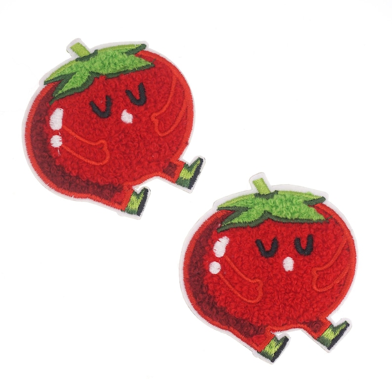 Puede incluir: Dos parches bordados en fieltro rojo de un tomate de dibujos animados con un tallo y hojas verdes. El tomate tiene una cara sonriente con dos ojos y una boca. El tomate tiene dos brazos y dos piernas peque&ntilde;as.