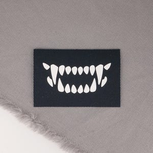 May include: A black rectangular patch featuring a white, stylized set of sharp teeth. The patch is set against a gray fabric background with a frayed edge. The design is simple and bold.
