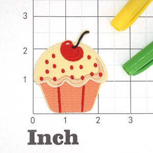 Może przedstawiać: Ż&oacute;łty cupcake z czerwonym i białym lukrem i czerwoną wiśnią na g&oacute;rze. Cupcake ma około 5 cm szerokości.