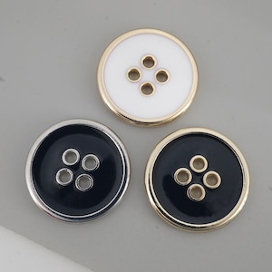 May include: Three circular buttons, each with four holes. One is white with a gold rim, another is black with a silver rim, and the third is black with a gold rim. They are set against a grey backdrop.