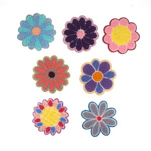 May include: Set of seven colorful felt flower patches. The flowers are in various colors, including blue, purple, pink, red, yellow, and gray. Each flower has a different design and is surrounded by a decorative border.