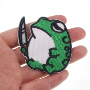 May include: Green embroidered frog holding a knife, a popular patch for jackets, bags, and other accessories.
