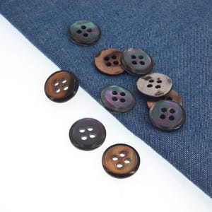 以下が含まれることがあります： それぞれ4つの穴がある、虹色のボタンのコレクション。ボタンの色は、茶色、黒、真珠のような光沢など様々です。一部のボタンは青いデニム生地の上に、その他は白い背景にあります。縫製プロジェクトに最適です。