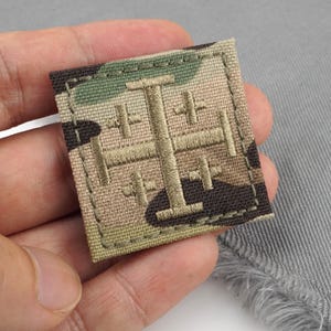 Puede incluir: Un parche cuadrado con un patrón de camuflaje y un diseño bordado en color beige. El parche tiene un borde cosido y se sostiene sobre un fondo de tela gris.