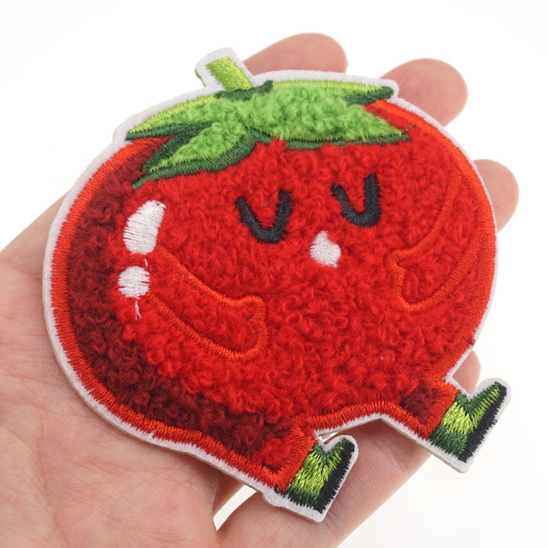 Puede incluir: Parche bordado de felpa roja en forma de tomate con una cara sonriente y hojas verdes. El tomate tiene ojos blancos y una peque&ntilde;a nariz blanca.