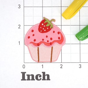 Może przedstawiać: R&oacute;żowy cupcake z czerwoną truskawką na wierzchu. Cupcake ma biały lukier z r&oacute;żowymi posypkami. Cupcake ma około 5 cm szerokości.