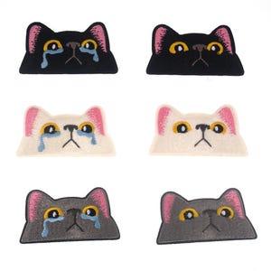 May include: Six embroidered iron-on patches of crying cats. The patches are in three colours: black, white, and grey. Each patch features a cat with pink ears and blue teardrops.