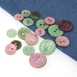 May include: A selection of decorative buttons in shades of pink, green, and brown. The buttons vary in size and feature a floral pattern. The background is a blue fabric. Measurements are not available.