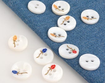 May include: A collection of white, round buttons with two holes each. The buttons are decorated with embroidered floral designs in shades of orange, red, and blue. The buttons are arranged on a blue fabric surface.