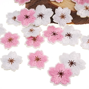 Może przedstawiać: Haftowane naszywki w kształcie kwiatów w kolorze białym i różowym, przypominające kwiaty wiśni. Naszywki mają szczegółowy wzór z brązowymi środkami i są rozrzucone na białej powierzchni. Idealne do dekoracji odzieży lub rękodzieła.