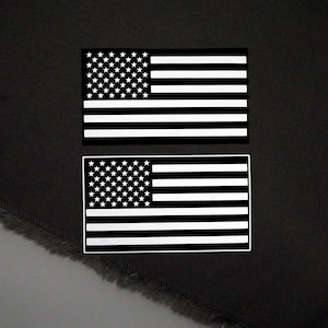 May include: Two black and white American flag patches. Each patch features a field of white stars in the upper left corner and alternating white and black stripes. The patches are rectangular and set against a dark background.