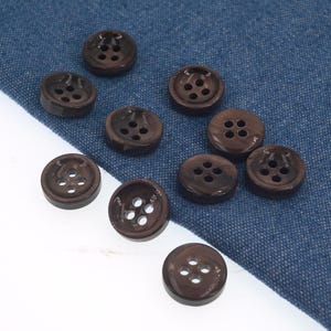 May include: A collection of dark brown, round buttons with four holes each. The buttons are arranged on a blue fabric background and a white surface. The buttons have a glossy finish and are made of a durable material.