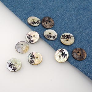 Może przedstawiać: Kolekcja okrągłych, dekoracyjnych guzików z kwiatowymi wzorami. Guziki są w różnych odcieniach bieli, brązu i opalizujących, z czarnymi wzorami kwiatowymi. Mają dwa lub cztery otwory do szycia i są ułożone na niebieskim tle z tkaniny.