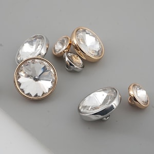 May include: A selection of decorative buttons featuring clear, faceted crystal centres. The buttons have gold and silver metal rims. The buttons vary in size and shape, with round and oval designs. The buttons are on a grey surface.