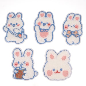 May include: Five white plush bunny patches with blue outlines. Each bunny has pink cheeks and embroidered details. One bunny is holding a carrot, one is holding a cup with a straw, one has a bow tie, and two are smiling.