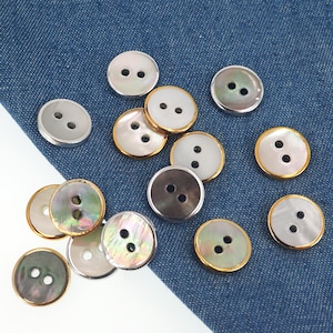 Op de afbeelding: Een verzameling ronde knopen in verschillende kleuren, waaronder zilver, goud en iriserend parelmoer. Elke knoop heeft twee gaten om te naaien. De knopen zijn verspreid op een blauwe denim stof.