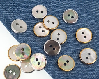 Könnte beinhalten: Eine Sammlung runder Knöpfe in verschiedenen Farben, darunter Silber, Gold und schillerndes Perlmutt. Jeder Knopf hat zwei Löcher zum Annähen. Die Knöpfe sind auf einem blauen Jeansstoff verstreut.