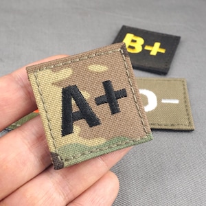 以下が含まれることがあります： 3つのグレードマーク付き刺繍パッチのクローズアップ。メインパッチは、迷彩柄の背景に黒い「A+」を表示しています。他のパッチは、黒地に黄色の「B+」とオリーブドラブ地に白の「D-」を表示しています。