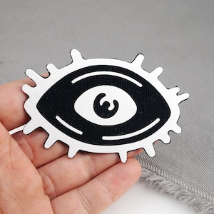 Op de afbeelding: Een zwart-wit oogvormige patch. Het oog heeft een zwart centrum met een witte iris en een witte omtrek. De patch wordt tegen een grijze achtergrond gehouden.