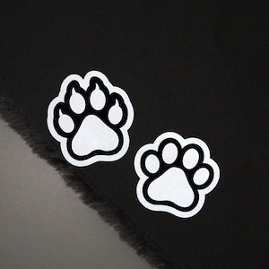 May include: Two white paw print stickers with black outlines are displayed against a black background. The stickers are in the shape of a dog's paw, with the pads and toes clearly defined. The stickers are likely for pet lovers.