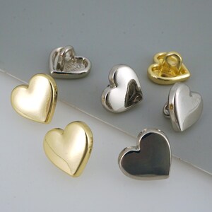 May include: An assortment of heart-shaped buttons in gold, silver, and a darker metallic finish. The buttons have a smooth, polished surface and a loop on the back for sewing. The buttons are suitable for various sewing projects.