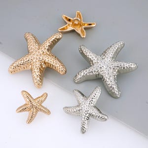 Op de afbeelding: Een verzameling metalen zeesterversieringen in goud en zilver. De zeesterren variëren in grootte en hebben een textuur. Deze decoratieve elementen zijn geschikt voor knutselprojecten.