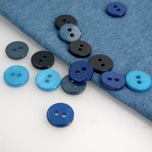 以下が含まれることがあります： さまざまな青と黒の色合いの丸いボタンのコレクション。 ボタンには2つの穴があり、デニム生地の上に散らばっています。 ボタンは、明るいターコイズから濃いネイビーまで、サイズと色合いが異なります。