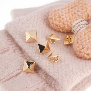 May include: Close-up of gold-colored pyramid-shaped studs and buttons with a black accent on a pink knitted fabric. Some buttons have a loop on the back. The image also shows a peach-colored knitted item.