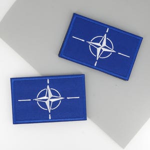 Pode incluir: Duas aplicações retangulares azuis com o símbolo da NATO bordado em branco. O símbolo apresenta uma rosa dos ventos branca dentro de um círculo branco, com linhas brancas. As aplicações são exibidas numa superfície cinzenta.