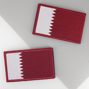 Puede incluir: Dos parches rectangulares con la bandera de Qatar. Cada parche tiene un fondo granate con un borde blanco dentado en el lado izquierdo. Los parches están bordeados con un ribete azul oscuro.