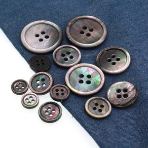May include: A collection of iridescent shell buttons in various sizes and shades of gray, black, and green. The buttons have four holes and are arranged on a blue fabric background. The buttons range in size from small to large.