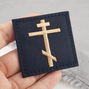 May include: A navy blue embroidered patch with a gold Orthodox cross design. The square patch has stitched edges. The cross features a horizontal bar, a slanted lower bar, and a smaller bar at the top.