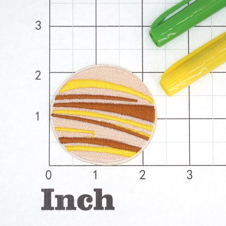 May include: A round, embroidered patch with a design of a planet with brown and yellow stripes on a pink background. The patch is approximately 1 inch in diameter.
