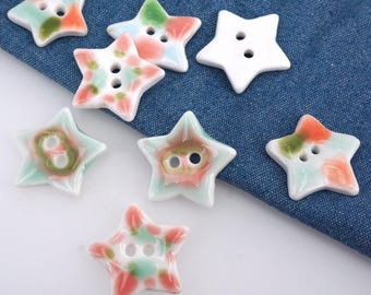 May include: A collection of star-shaped ceramic buttons in various colors. The buttons feature a glossy finish and are painted with shades of green, orange, and pink. Some buttons are solid white. Each button has two holes for sewing.