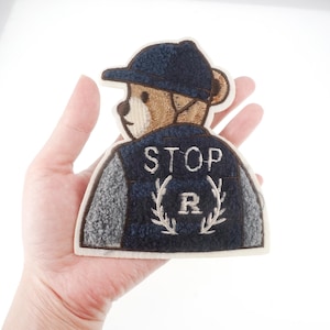 May include: Embroidered iron-on patch featuring a brown teddy bear wearing a blue and gray jacket with a white baseball cap. The jacket has the text "STOP" and a wreath with the letter "R" embroidered on the back.