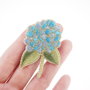 Op de afbeelding: Een geborduurde hortensia bloemenpatch in blauw en goud. De bloem is gedetailleerd met lichtblauwe en donkerblauwe bloemblaadjes en gouden accenten. De bloem is bevestigd aan een groene stengel met twee bladeren.