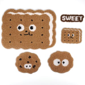 Puede incluir: Cinco parches de fieltro con forma de galletas. Los parches son marrones con detalles blancos y tienen caras lindas. Un parche dice "SWEET" en letras de fieltro marrones.